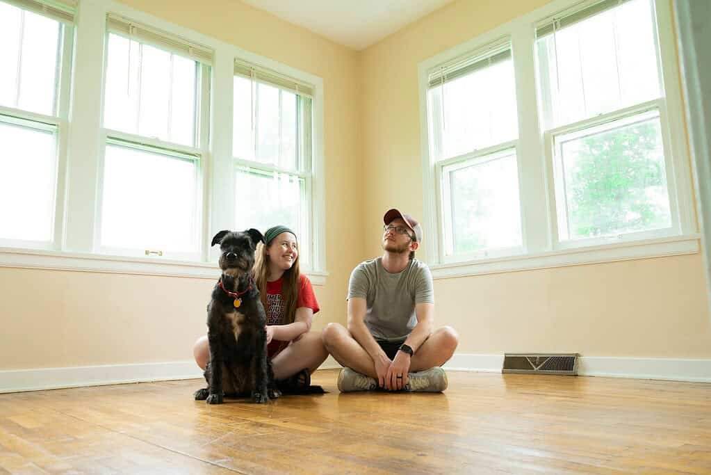 Dog with owner and friend sitting on hardwood floor in bright room with large windows.