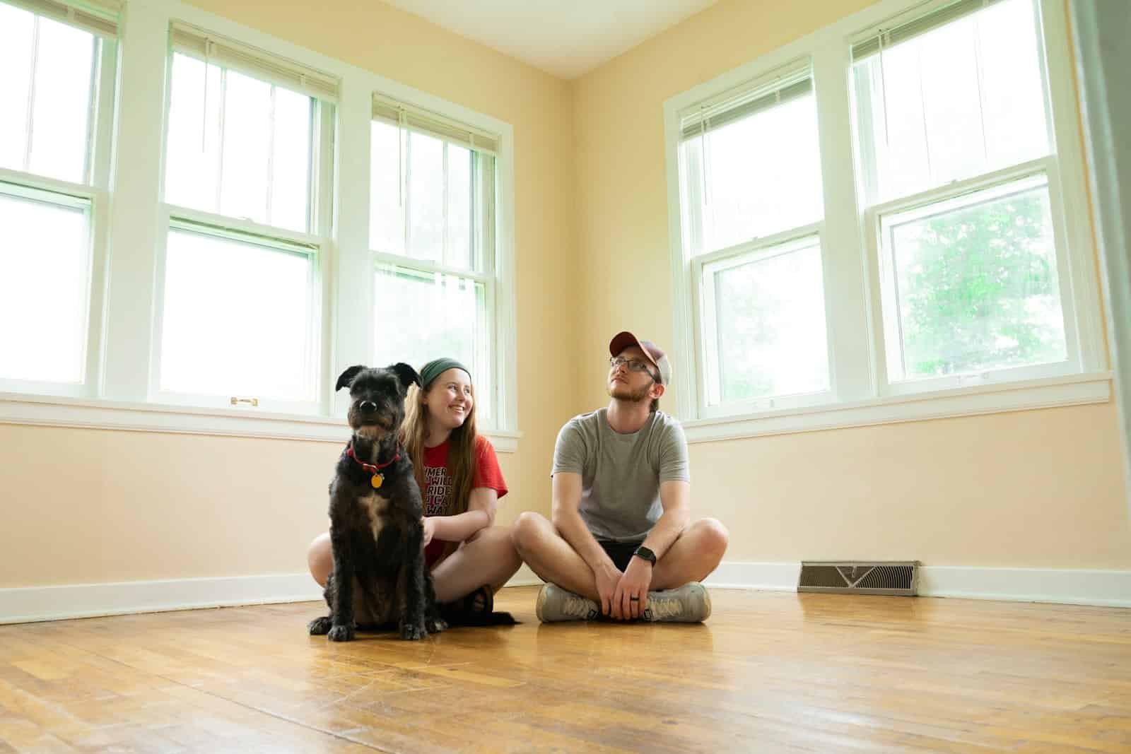 Dog with owner and friend sitting on hardwood floor in bright room with large windows.
