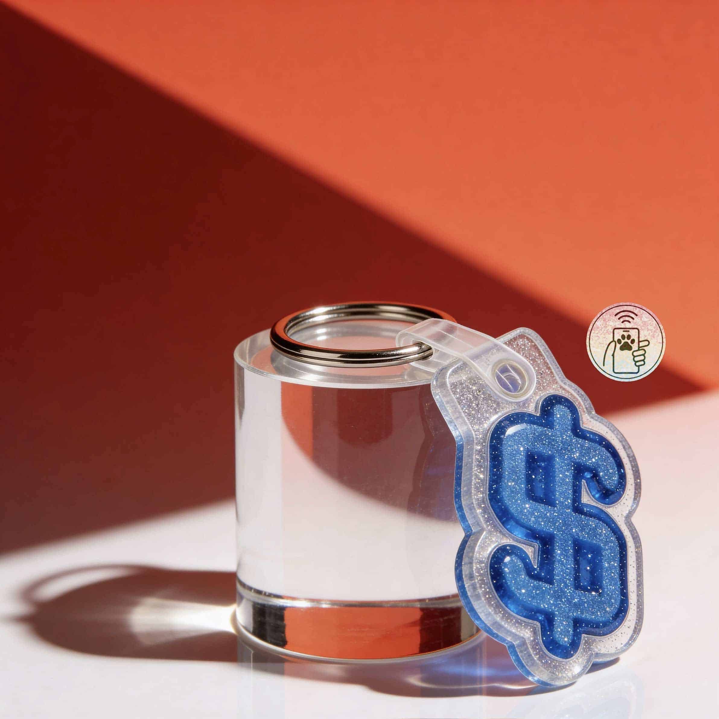 Clear acrylic NFC pet ID tag shaped like a dollar sign with glitter, attached to a metal keyring — Shiloh's House smart NFC pet tag with free companion Lohji app.