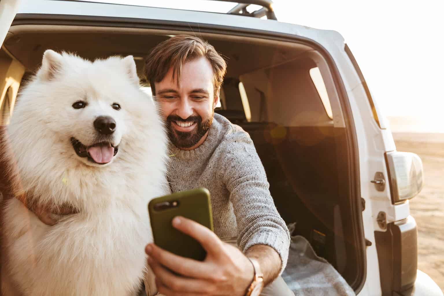Man and fluffy dog looking at smartphone in car trunk.