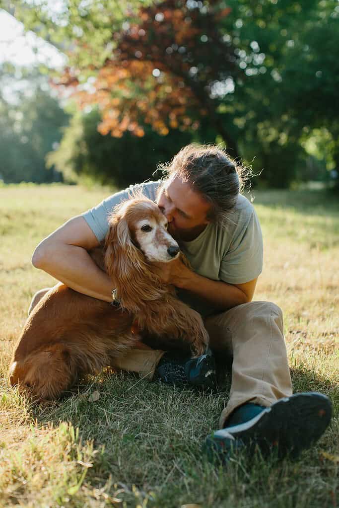 Woman hugging her dog in a sunny park with trees in the background.