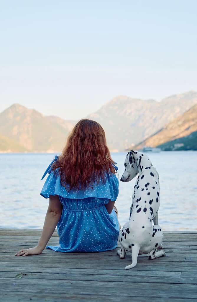 Girl with long red hair sitting with Dalmatian dog on dock by lake.
