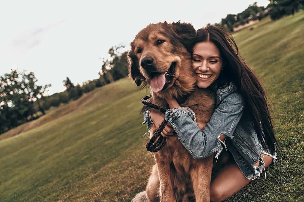 Smiling woman hugging a large long-haired golden-brown dog on a grassy hillside